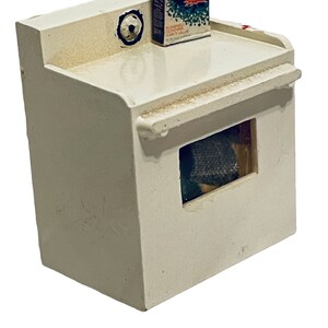 Peut inclure: Une petite machine &agrave; laver blanche avec une petite bo&icirc;te de d&eacute;tergent "Rinso" sur le dessus. La machine &agrave; laver a une ouverture rectangulaire &agrave; l'avant et un motif circulaire en haut &agrave; gauche. La bo&icirc;te de d&eacute;tergent est rectangulaire avec du texte et des graphiques.