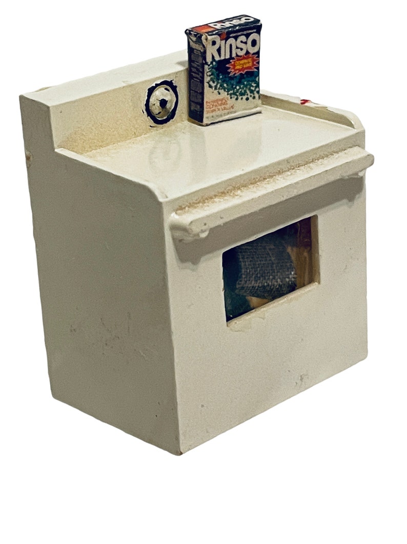 Peut inclure: Une petite machine &agrave; laver blanche avec une petite bo&icirc;te de d&eacute;tergent "Rinso" sur le dessus. La machine &agrave; laver a une ouverture rectangulaire &agrave; l'avant et un motif circulaire en haut &agrave; gauche. La bo&icirc;te de d&eacute;tergent est rectangulaire avec du texte et des graphiques.
