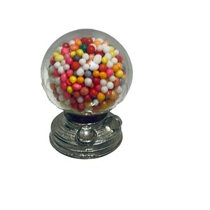 May include: A miniature gumball machine with a clear, round globe filled with colorful gumballs. The base is silver-colored with a classic design. A small, nostalgic candy dispenser.