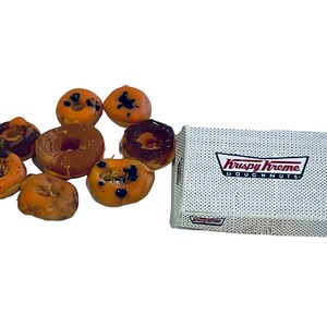 May include: A box of orange and brown glazed donuts with black icing and sprinkles. The box is white with the words "Krispy Kreme Doughnuts" printed in red and black.