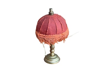 May include: Miniature table lamp with a burgundy fabric shade and a gold-toned base. The shade has a pleated design and is trimmed with fringe. The lamp is a decorative item, likely for a dollhouse or small display.