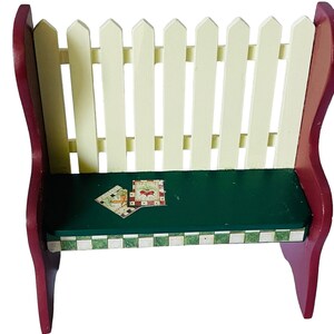 May include: A small wooden bench with a white picket fence back, a green top, and a checkered border. The bench has a red base and is decorated with a small, colourful patchwork design.