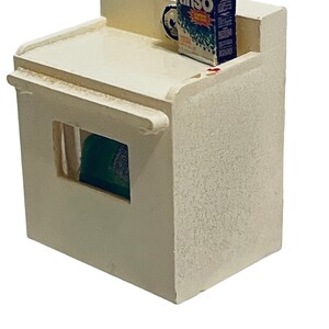 Peut inclure: Une maquette miniature de machine &agrave; laver blanc cass&eacute; avec une petite bo&icirc;te de d&eacute;tergent "Rinso" sur le dessus. La bo&icirc;te est principalement bleue, verte et rouge. Un objet vert est visible &agrave; travers une ouverture rectangulaire &agrave; l'avant.