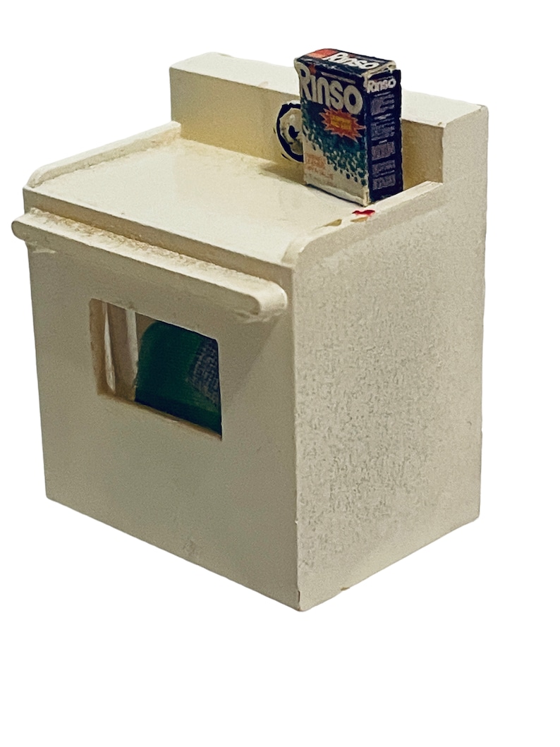 Peut inclure: Une maquette miniature de machine &agrave; laver blanc cass&eacute; avec une petite bo&icirc;te de d&eacute;tergent "Rinso" sur le dessus. La bo&icirc;te est principalement bleue, verte et rouge. Un objet vert est visible &agrave; travers une ouverture rectangulaire &agrave; l'avant.