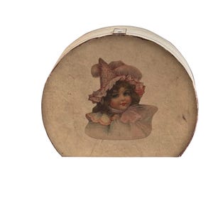 Peut inclure: Une boîte à chapeau ronde et vintage avec un extérieur crème. Le couvercle présente une charmante illustration d'un jeune enfant portant un bonnet et une robe roses. La boîte est probablement en carton et montre des signes d'âge.