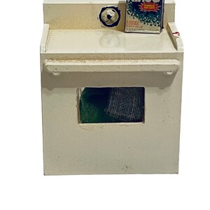 Peut inclure: Une petite machine &agrave; laver blanche avec une petite bo&icirc;te de d&eacute;tergent "Rinso" sur le dessus. La machine &agrave; laver a une ouverture rectangulaire r&eacute;v&eacute;lant du tissu vert et gris &agrave; l'int&eacute;rieur. La bo&icirc;te de d&eacute;tergent a du texte rouge, blanc et bleu.
