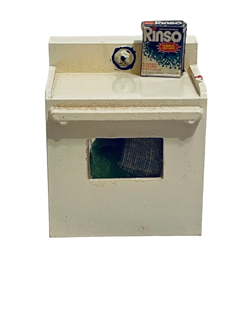 Peut inclure: Une petite machine &agrave; laver blanche avec une petite bo&icirc;te de d&eacute;tergent "Rinso" sur le dessus. La machine &agrave; laver a une ouverture rectangulaire r&eacute;v&eacute;lant du tissu vert et gris &agrave; l'int&eacute;rieur. La bo&icirc;te de d&eacute;tergent a du texte rouge, blanc et bleu.