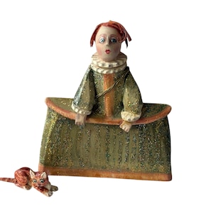 May include: A whimsical figurine of a person with red hair and a ruffled collar, wearing a green dress with gold accents. A small, orange and brown cat figurine is lying down nearby. The figurine has a glittery finish.