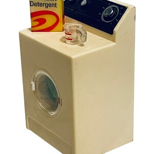 Peut inclure: Une machine &agrave; laver jouet miniature de couleur cr&egrave;me avec une porte transparente. Une bo&icirc;te jaune de "LAUNDRY Detergent" est pos&eacute;e sur le dessus, ainsi qu'un petit verre doseur. Le panneau de commande est noir avec un cadran et deux boutons. Un jouet amusant de style vintage.