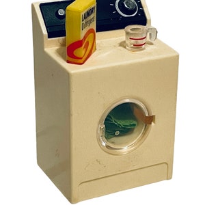 Peut inclure: Une machine &agrave; laver jouet vintage de couleur cr&egrave;me avec un panneau de commande noir. Une bo&icirc;te jaune de "LAUNDRY Detergent" et un petit verre doseur sont pos&eacute;s dessus. La porte de la machine est transparente, r&eacute;v&eacute;lant du tissu vert &agrave; l'int&eacute;rieur.