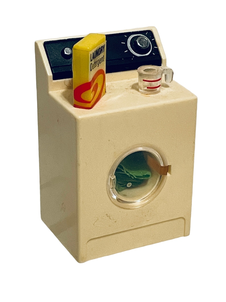 Peut inclure: Une machine &agrave; laver jouet vintage de couleur cr&egrave;me avec un panneau de commande noir. Une bo&icirc;te jaune de "LAUNDRY Detergent" et un petit verre doseur sont pos&eacute;s dessus. La porte de la machine est transparente, r&eacute;v&eacute;lant du tissu vert &agrave; l'int&eacute;rieur.