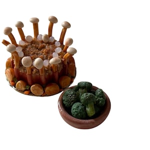 May include: A miniature food arrangement featuring a round dish with a meat-like filling, surrounded by what appears to be bacon and mushrooms. A small bowl of green broccoli is next to the dish.