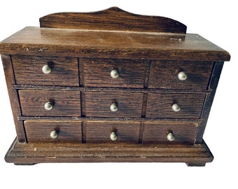 May include: A small wooden dresser with nine drawers, each with a silver knob. The dresser is made of dark wood and has a slightly distressed finish.