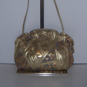 May include: A gold-colored leather shoulder bag with a patchwork design. The bag has a long strap and is hanging from a metal stand.