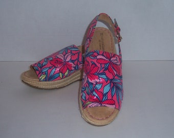 May include: A pair of pink floral wedge sandals with a buckle closure. The sandals have a jute sole and a pink floral print.