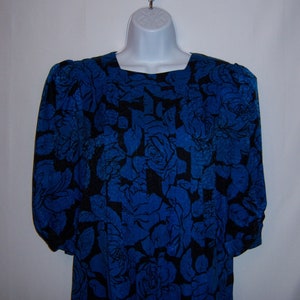 May include: A blue dress with a black floral pattern. The dress has a square neckline and long sleeves with gathered details at the shoulders and cuffs. The fabric is a vibrant blue with a contrasting black rose design.