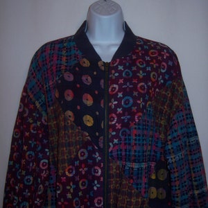 May include: A colorful patchwork bomber jacket with a zipper closure. The jacket features a variety of patterns, including circles, flowers, and plaid, in shades of blue, purple, pink, and yellow. The collar and trim are a solid navy blue.