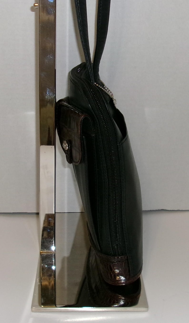 May include: Black leather handbag with a brown crocodile-textured accent pocket and trim. The bag has a long black strap and a silver zipper. The bag is displayed on a silver stand.