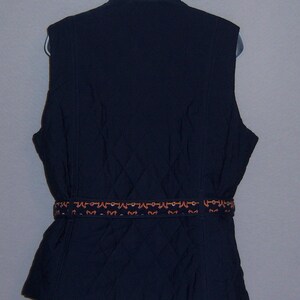 May include: A navy blue quilted vest with a decorative brown and orange belt. The vest has a stand-up collar and a zipper closure.