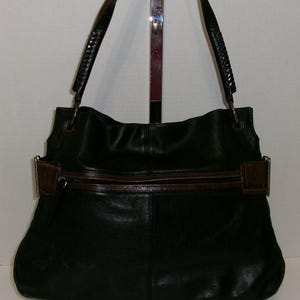 K&ouml;nnte beinhalten: Schwarze Leder-Umh&auml;ngetasche mit braunen Akzenten. Die Tasche hat einen Rei&szlig;verschluss, einen braunen Lederriemen und geflochtene Griffe. Die Tasche wird vor einem neutralen Hintergrund gezeigt.