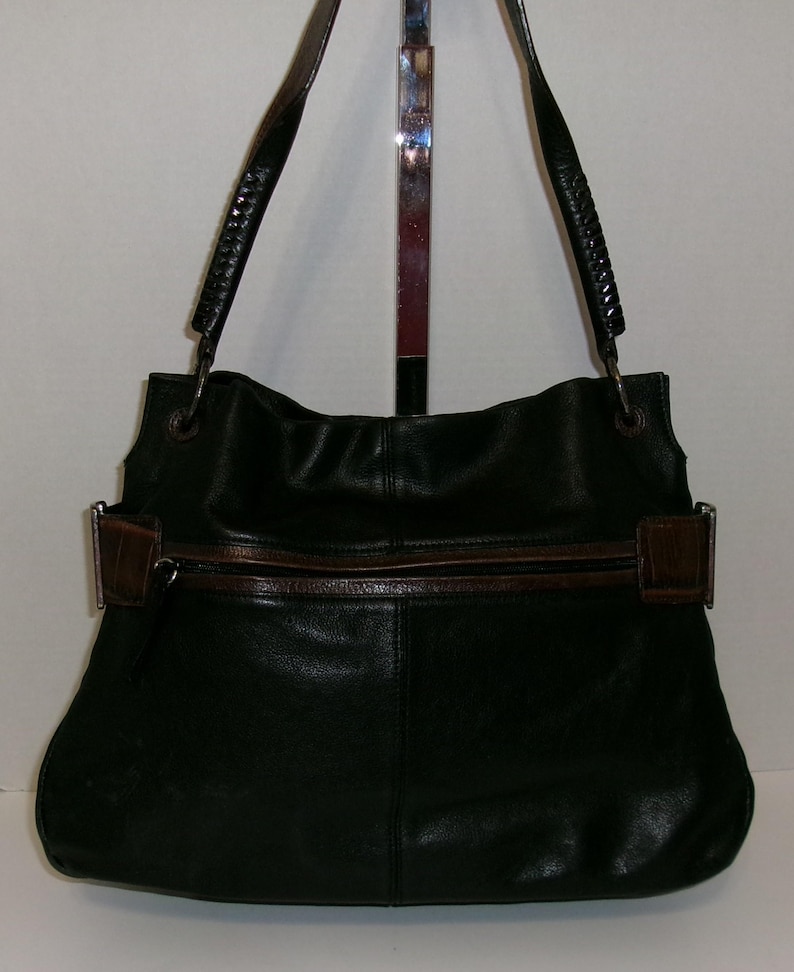 K&ouml;nnte beinhalten: Schwarze Leder-Umh&auml;ngetasche mit braunen Akzenten. Die Tasche hat einen Rei&szlig;verschluss, einen braunen Lederriemen und geflochtene Griffe. Die Tasche wird vor einem neutralen Hintergrund gezeigt.