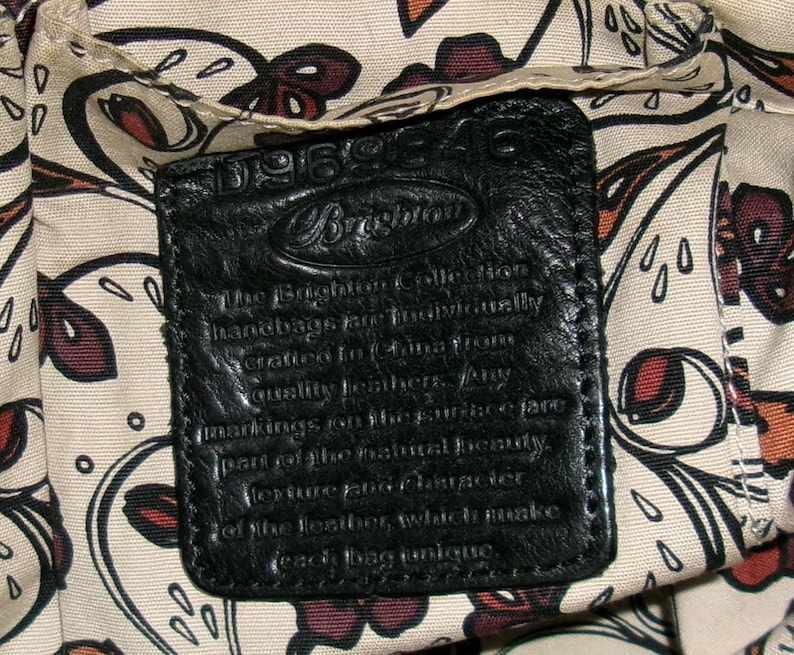 K&ouml;nnte beinhalten: Nahaufnahme eines schwarzen Lederetiketts mit gepr&auml;gtem Text, wahrscheinlich von einer Handtasche. Das Etikett ist rechteckig mit abgerundeten Ecken und zeigt das Brighton-Logo. Der Hintergrund zeigt einen gemusterten Stoff mit floralen und abstrakten Designs.