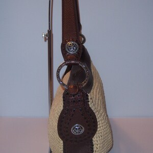 Może przedstawiać: Brązowa sk&oacute;rzana torebka z kremowym haftem, srebrnymi okuciami i kwiatowym wzorem. Torebka znajduje się na stojaku wystawowym.