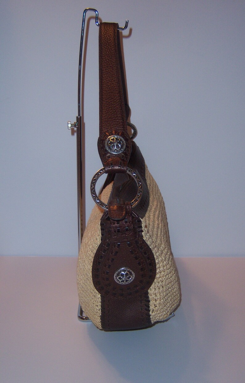 Może przedstawiać: Brązowa sk&oacute;rzana torebka z kremowym haftem, srebrnymi okuciami i kwiatowym wzorem. Torebka znajduje się na stojaku wystawowym.