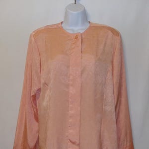 Puede incluir: Una blusa de manga larga de color melocotón con un sutil patrón geométrico. La blusa tiene un cierre de botones en la parte delantera y un cuello mandarín.
