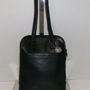 May include: Black leather tote bag with a silver-tone charm. The bag has a rectangular shape with a rounded top, a zippered closure, and two long shoulder straps. The bag is displayed on a silver stand.