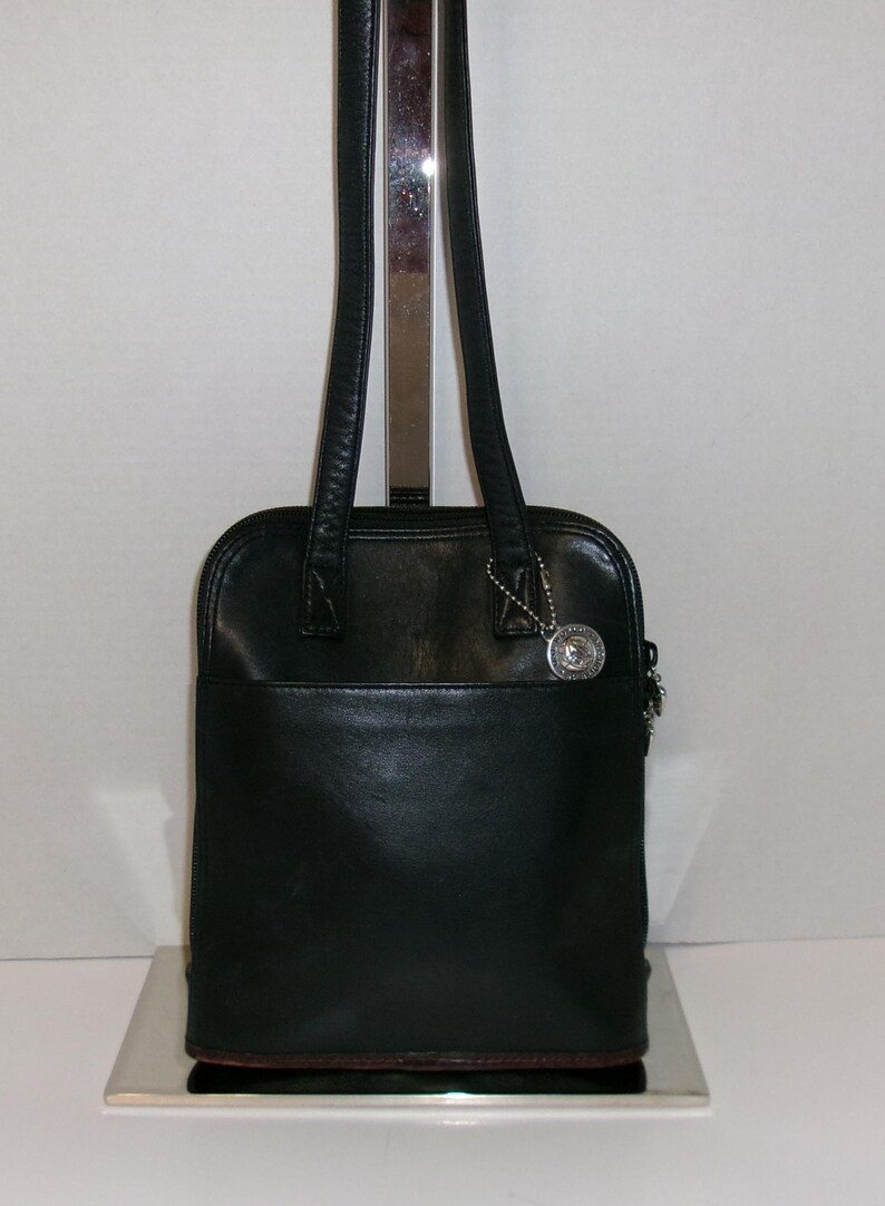 May include: Black leather tote bag with a silver-tone charm. The bag has a rectangular shape with a rounded top, a zippered closure, and two long shoulder straps. The bag is displayed on a silver stand.