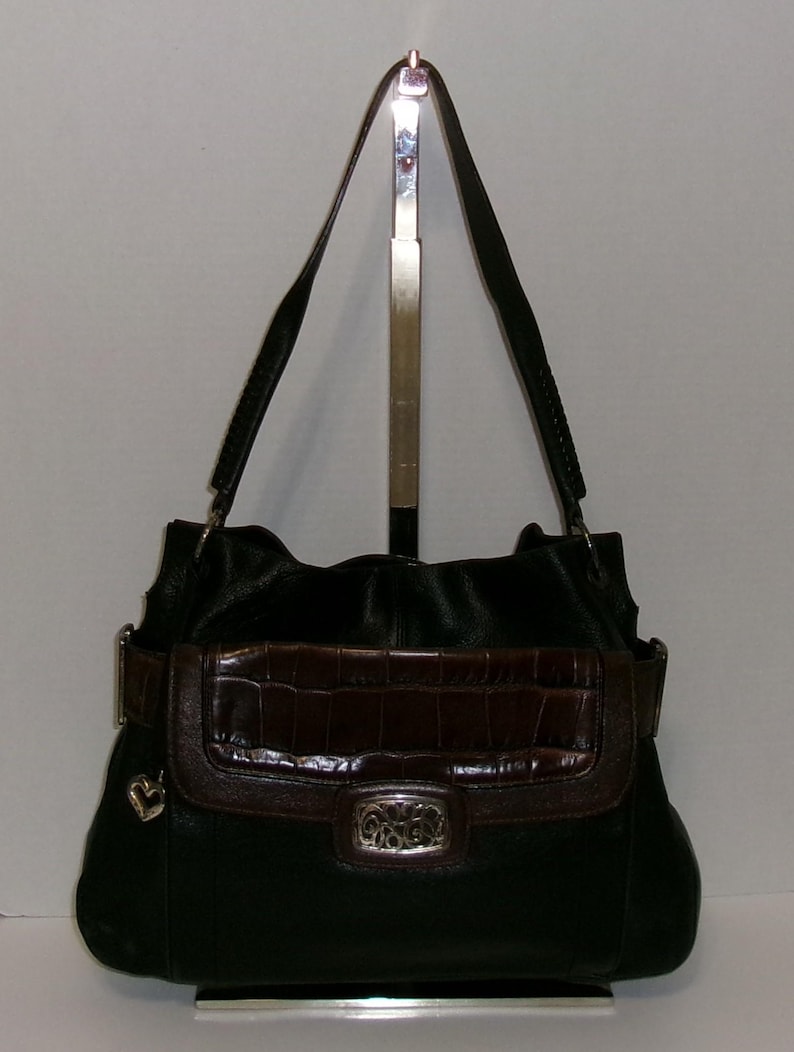 K&ouml;nnte beinhalten: Schwarze Leder-Umh&auml;ngetasche mit braunem, krokodiltexturiertem &Uuml;berschlag und Riemen. Die Tasche hat eine silberfarbene Schnalle und einen dekorativen Herzanh&auml;nger. Der Riemen ist verstellbar und die Tasche wird auf einem silbernen St&auml;nder pr&auml;sentiert.