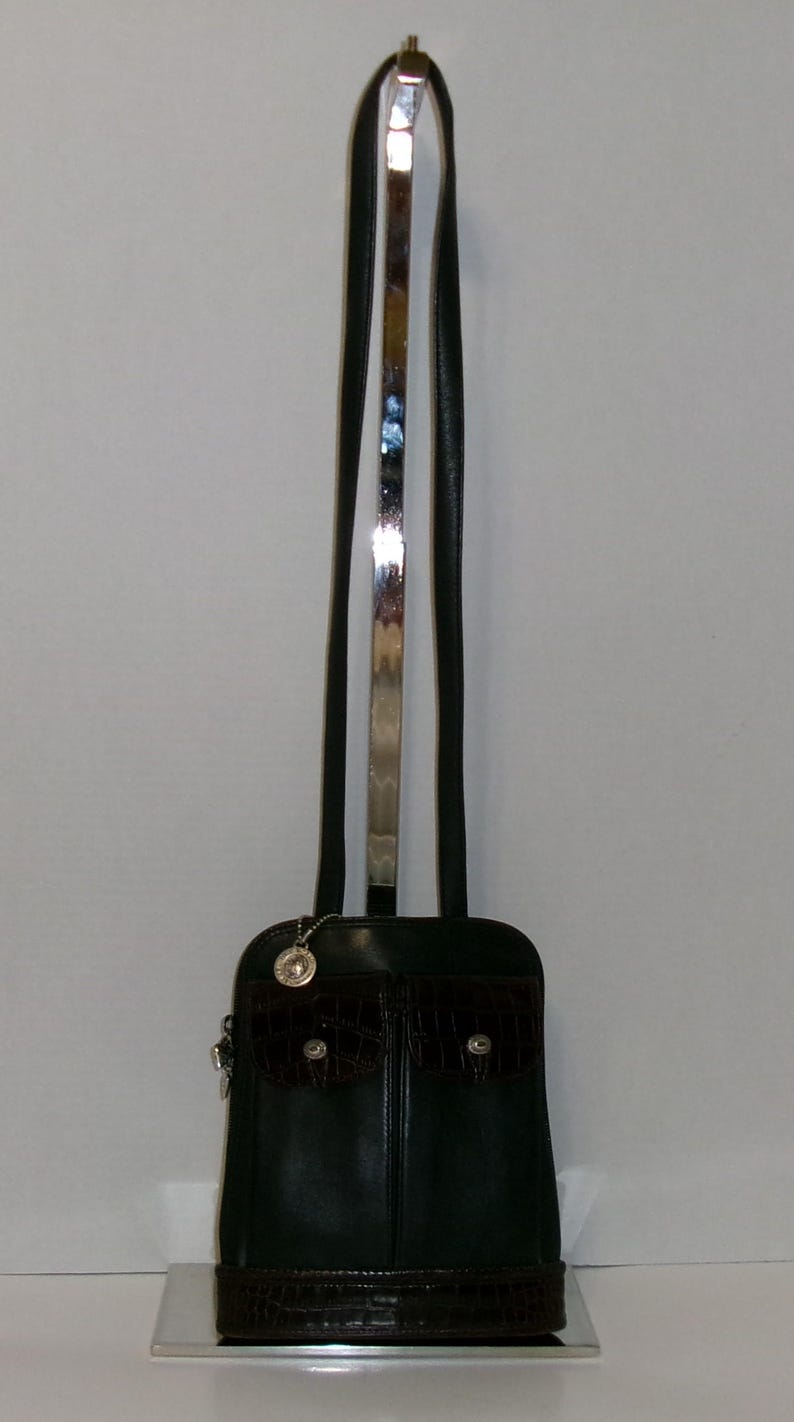 May include: A black leather handbag with a dark brown crocodile-textured front pocket and base. The bag has a long, thin strap with a silver metal accent. A small silver charm hangs from the top of the bag.