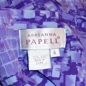 Puede incluir: Una etiqueta blanca con el texto "Adrianna Papell" en letras rojas. La etiqueta tambi&eacute;n tiene una etiqueta de talla 8 y una etiqueta m&aacute;s peque&ntilde;a que dice "100% Seda Hecho en China".
