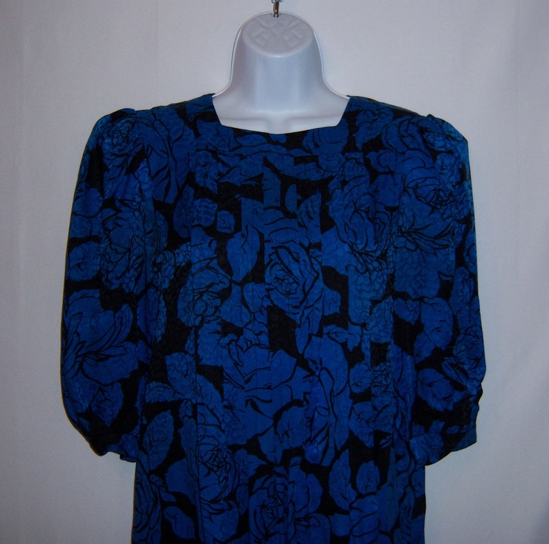 May include: A blue dress with a black floral pattern. The dress has a square neckline and long sleeves with gathered details at the shoulders and cuffs. The fabric is a vibrant blue with a contrasting black rose design.