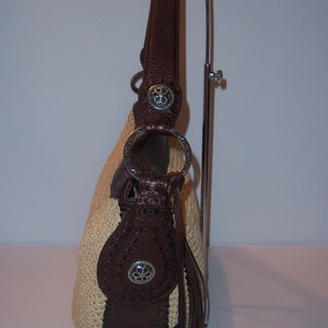 Może przedstawiać: Brązowa sk&oacute;rzana torebka ze słomy z srebrnym serduszkiem i srebrną klamrą. Torebka ma brązowy sk&oacute;rzany pasek i frędzle.