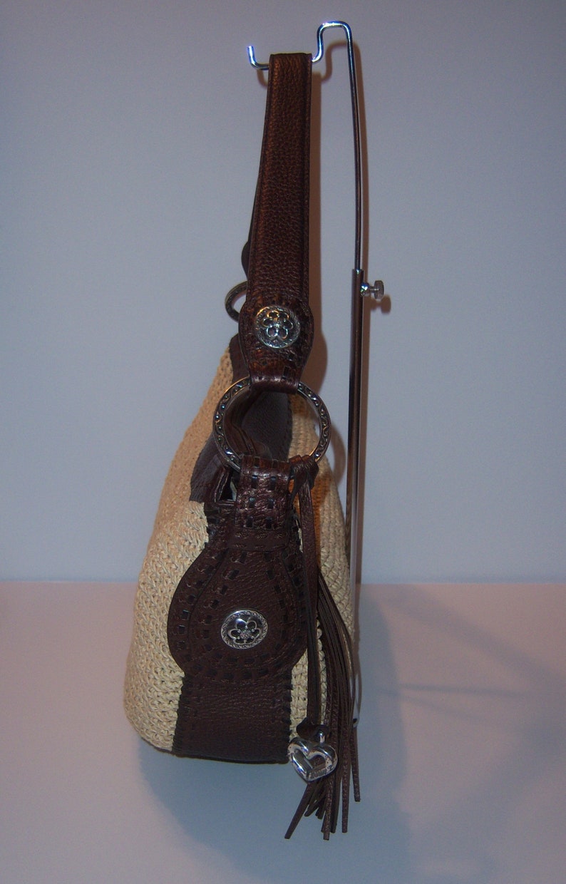 Może przedstawiać: Brązowa sk&oacute;rzana torebka ze słomy z srebrnym serduszkiem i srebrną klamrą. Torebka ma brązowy sk&oacute;rzany pasek i frędzle.