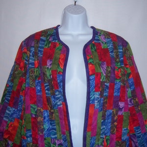 May include: A colorful, open-front jacket with a patchwork design. The jacket features vertical stripes of red, blue, green, and purple fabric with floral patterns. A deep purple trim outlines the edges. The jacket is displayed on a white mannequin.