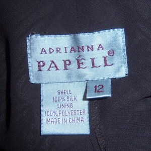 May include: A blue and white clothing label with the text "Adrianna Papell" and a size 12 tag. The label also includes the fabric content: "Shell 100% Silk, Lining 100% Polyester, Made in China."
