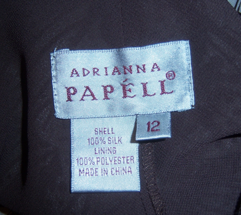 May include: A blue and white clothing label with the text "Adrianna Papell" and a size 12 tag. The label also includes the fabric content: "Shell 100% Silk, Lining 100% Polyester, Made in China."