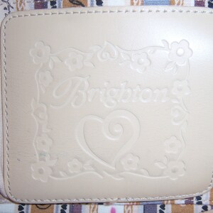 Może przedstawiać: Zbliżenie na kremową sk&oacute;rzaną łatę z wygrawerowanym słowem "Brighton" w ozdobnym, pisanym stylu. Słowo jest otoczone ozdobną ramką z kwiat&oacute;w i liści. W środku łatki znajduje się serce z ozdobnym wzorem w postaci spirali.