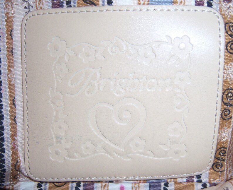 Może przedstawiać: Zbliżenie na kremową sk&oacute;rzaną łatę z wygrawerowanym słowem "Brighton" w ozdobnym, pisanym stylu. Słowo jest otoczone ozdobną ramką z kwiat&oacute;w i liści. W środku łatki znajduje się serce z ozdobnym wzorem w postaci spirali.