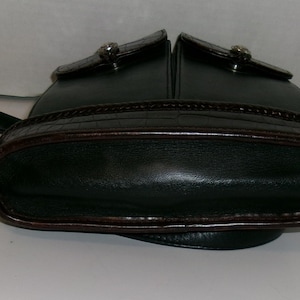 May include: Dark green leather handbag with a rounded bottom and two top compartments. The bag features a brown, textured trim and a silver-toned clasp on each compartment. A black strap is attached to the side.