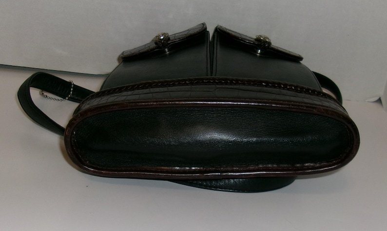 May include: Dark green leather handbag with a rounded bottom and two top compartments. The bag features a brown, textured trim and a silver-toned clasp on each compartment. A black strap is attached to the side.