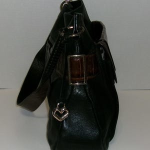 K&ouml;nnte beinhalten: Schwarze Leder-Umh&auml;ngetasche mit braunem Akzent und einem herzf&ouml;rmigen Charm in Silber. Die Tasche hat einen langen Riemen und einen Klappenverschluss mit braunem Lederakzent. Die Tasche ist aus strukturiertem Leder gefertigt.