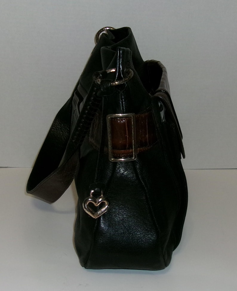 K&ouml;nnte beinhalten: Schwarze Leder-Umh&auml;ngetasche mit braunem Akzent und einem herzf&ouml;rmigen Charm in Silber. Die Tasche hat einen langen Riemen und einen Klappenverschluss mit braunem Lederakzent. Die Tasche ist aus strukturiertem Leder gefertigt.