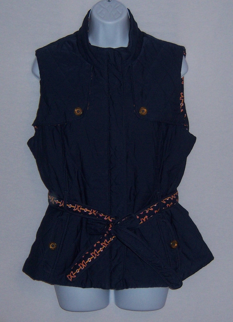 May include: A navy blue sleeveless vest with a zipper closure and a brown belt with a decorative pattern. The vest has two pockets on the front and a high collar.