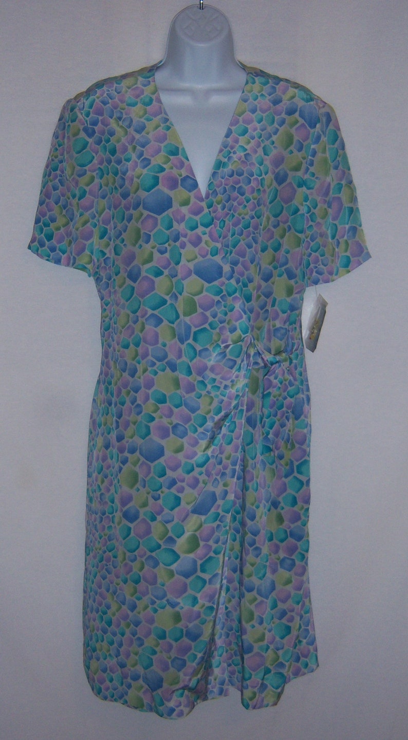 May include: A short-sleeved dress with a V-neck and a wrap-style design. The dress features a pattern of blue, green, and purple geometric shapes. The dress is displayed on a white mannequin.