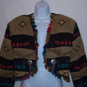 May include: A cropped jacket with a geometric pattern in black, tan, and red. The jacket has fringe trim in multiple colors and silver-tone button closures. The design includes circular and linear patterns.