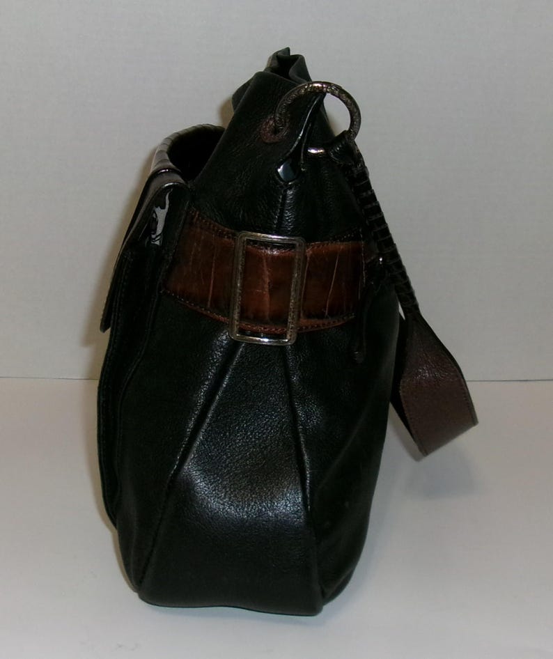 K&ouml;nnte beinhalten: Schwarze Lederhandtasche mit braunem, krokodiltexturiertem Akzentriemen und silberner Schnalle. Die Tasche hat einen Schulterriemen und einen Klappenverschluss. Die Tasche ist dunkelgr&uuml;n.